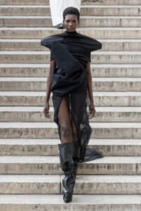 Model walking down stone steps in an avant-garde black dress with sculptural draping and tall black boots.