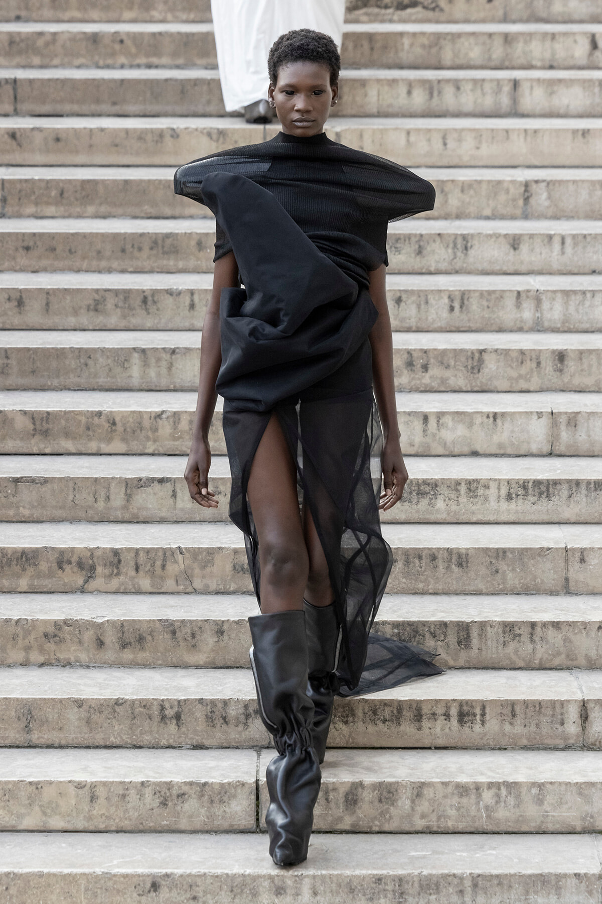 Model walking down stone steps in an avant-garde black dress with sculptural draping and tall black boots.
