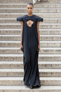 Model in a long, black gown with a circular chest cutout and sculptural shoulders standing on stone steps while walking down.
