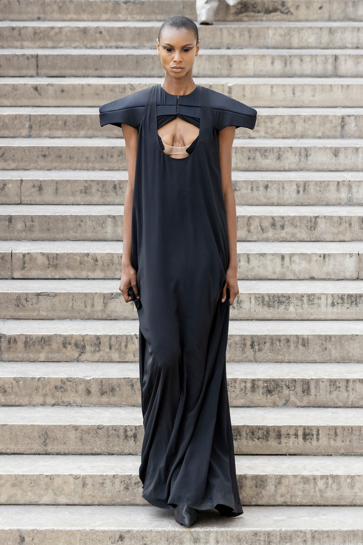 Model in a long, black gown with a circular chest cutout and sculptural shoulders standing on stone steps while walking down.
