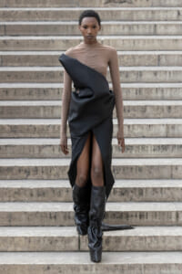Model walking down stone steps in a tan mesh top and a black draped gown with a high front slit, paired with tall black boots.