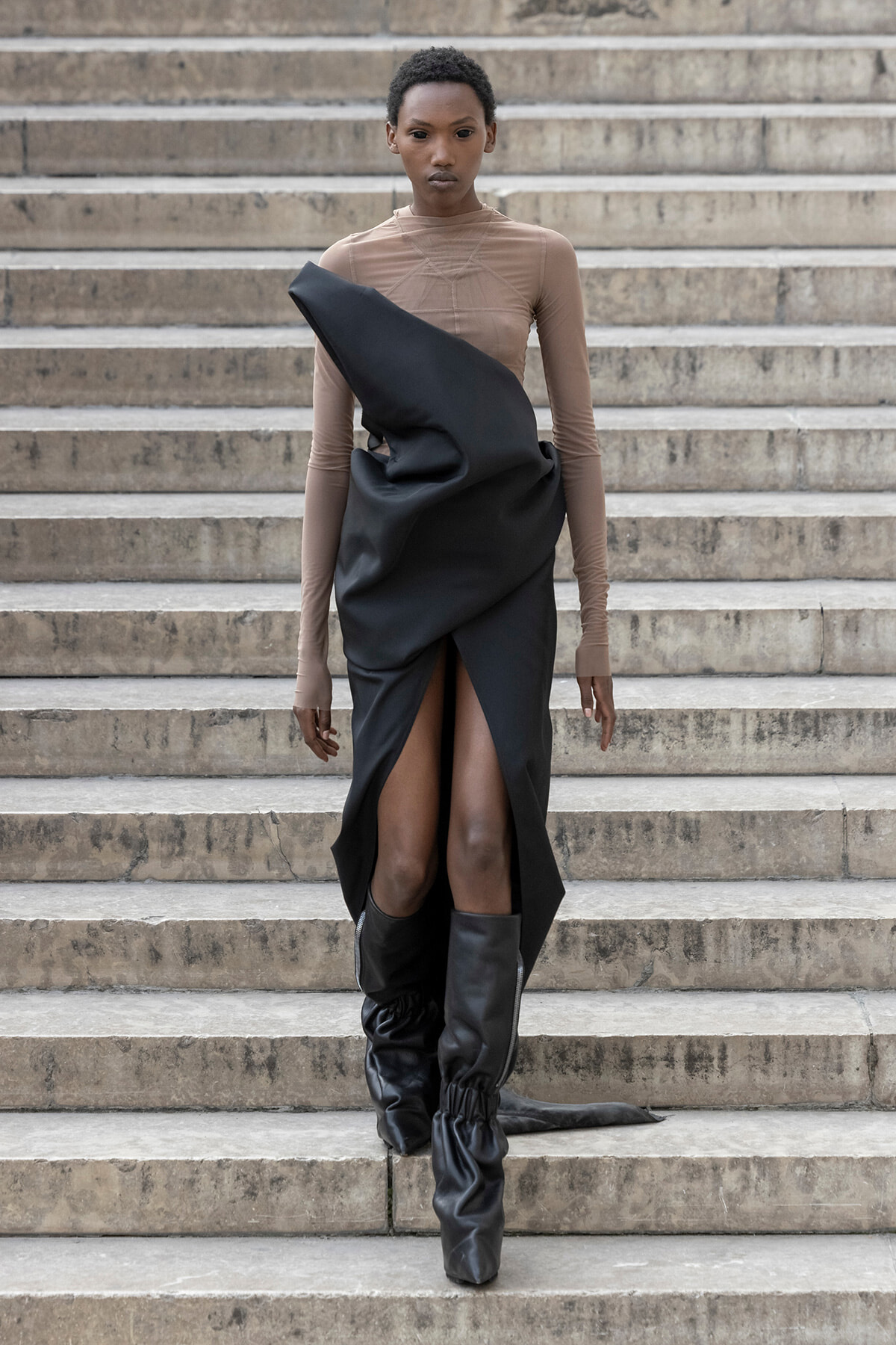 Model walking down stone steps in a tan mesh top and a black draped gown with a high front slit, paired with tall black boots.
