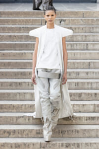 Avant-garde white cape over a white tunic and layered gray pants worn by a model descending stone steps at a fashion show.