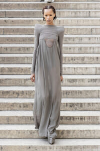 Model in a flowing, gray floor-length dress with a sheer panel, standing on concrete stairs on outdoor steps.