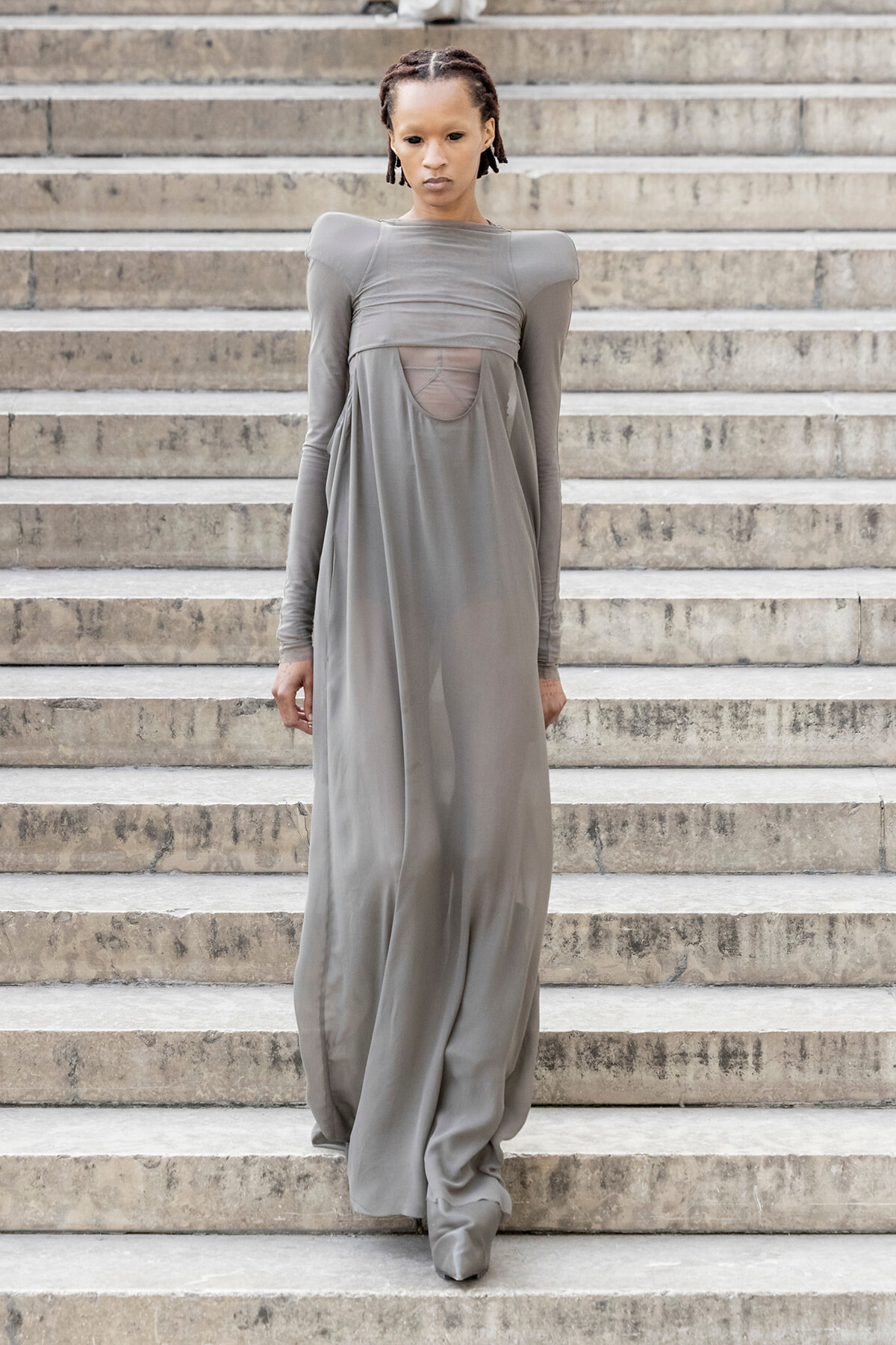 Model in a flowing, gray floor-length dress with a sheer panel, standing on concrete stairs on outdoor steps.