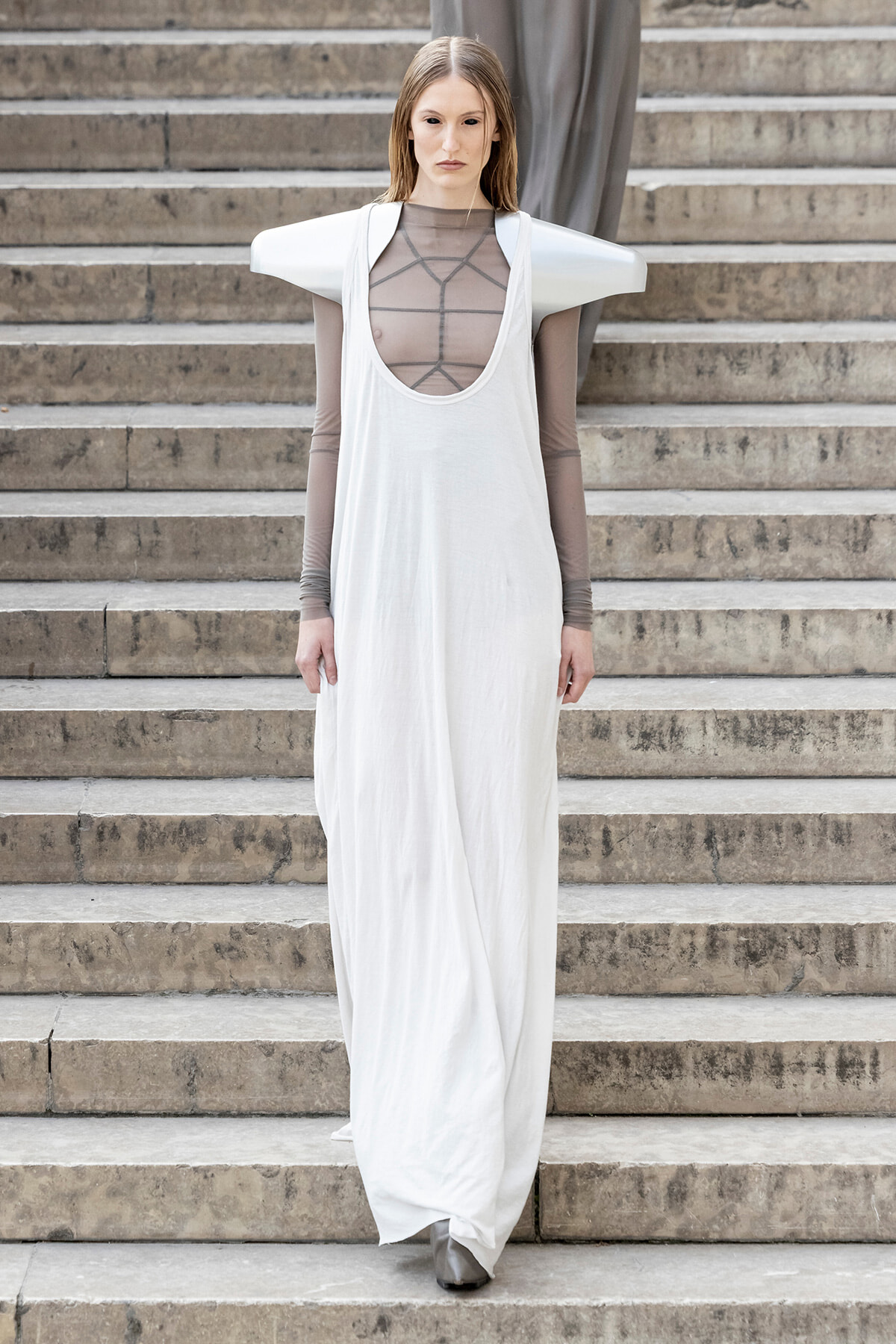 Model standing on stone steps, wearing a long white sleeveless dress with oversized shoulder armor and a sheer gray mesh top underneath.