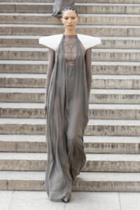 Model walking down stone stairs in a flowing gray gown with dramatic white sculptural shoulders on a runway look.