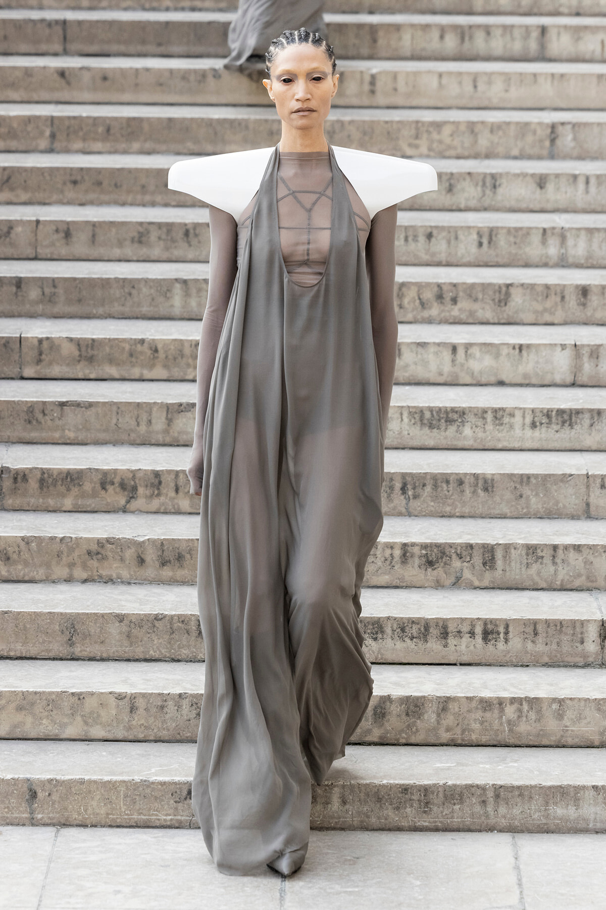 Model walking down stone stairs in a flowing gray gown with dramatic white sculptural shoulders on a runway look.