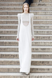 Woman in a long white gown with a deep scoop neckline standing on stone steps, facing the camera.