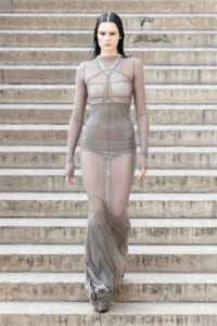 Model wearing a long sheer taupe gown walking down stone steps, hair slicked back.