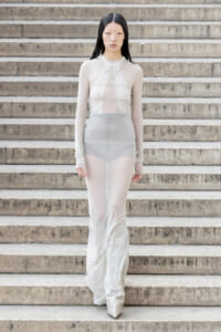Model walking down a concrete staircase in a sheer ivory full-length dress with long sleeves.