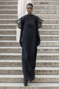 Model walking down stone steps in a black avant-garde gown with sheer mesh bodice and sculptural, layered ruffled sleeves.