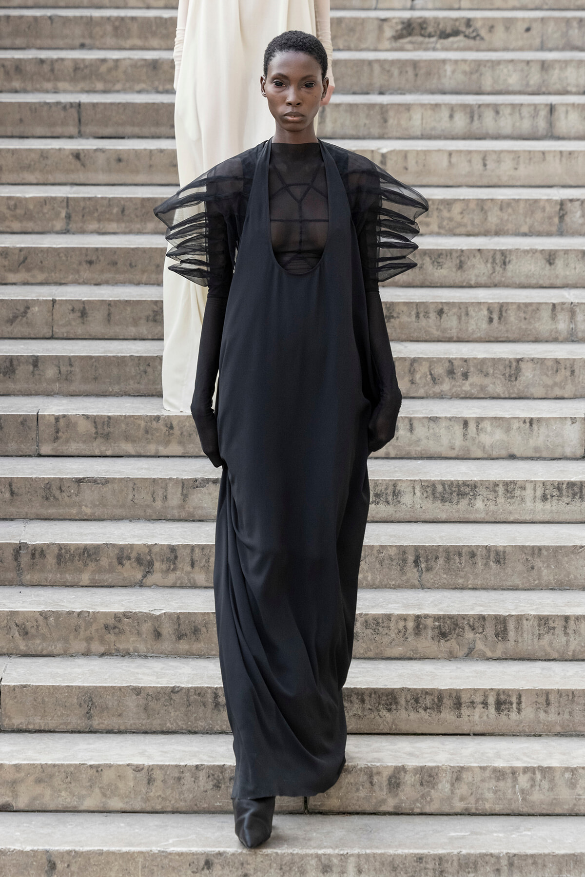 Model walking down stone steps in a black avant-garde gown with sheer mesh bodice and sculptural, layered ruffled sleeves.