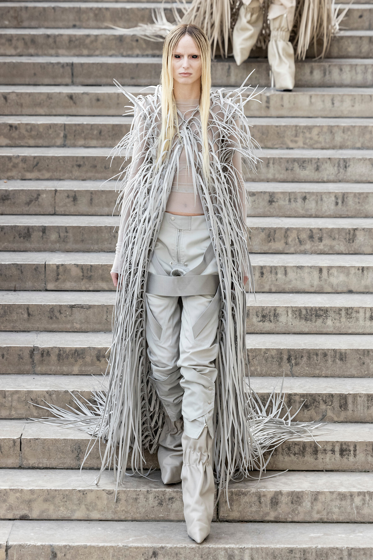 Model in a dramatic gray fringe coat with long shredded strands, walking down stone steps in a light-gray outfit and boots.
