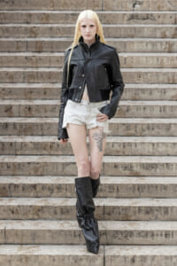 Young woman with long blonde hair in a black leather jacket, white shorts, and tall black boots standing on stone steps, facing the camera with a tattoo on her left thigh.