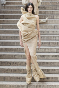 Model walking down stone steps in a beige sequined one-shoulder gown with a high slit and matching boots.