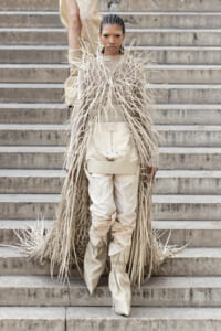 Model wearing a beige fringed coat with long, thin strands over a matching bodysuit on stone steps.