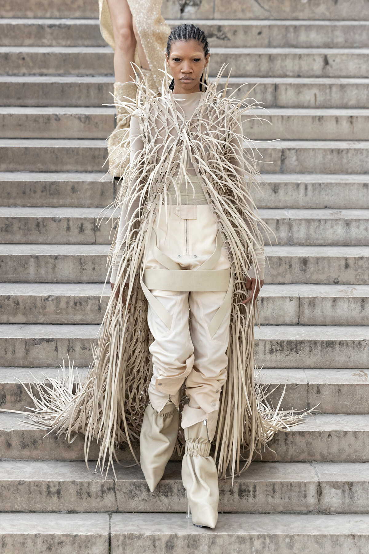 Model wearing a beige fringed coat with long, thin strands over a matching bodysuit on stone steps.