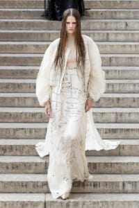 Model in a cream oversized jacket and dress covered with black diagram-like markings walking down stone steps.
