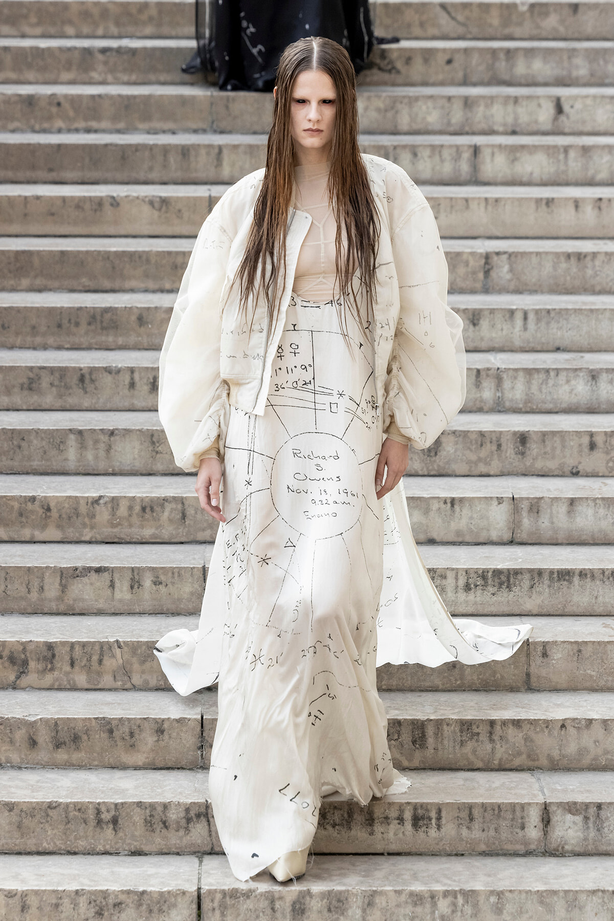 Model in a cream oversized jacket and dress covered with black diagram-like markings walking down stone steps.