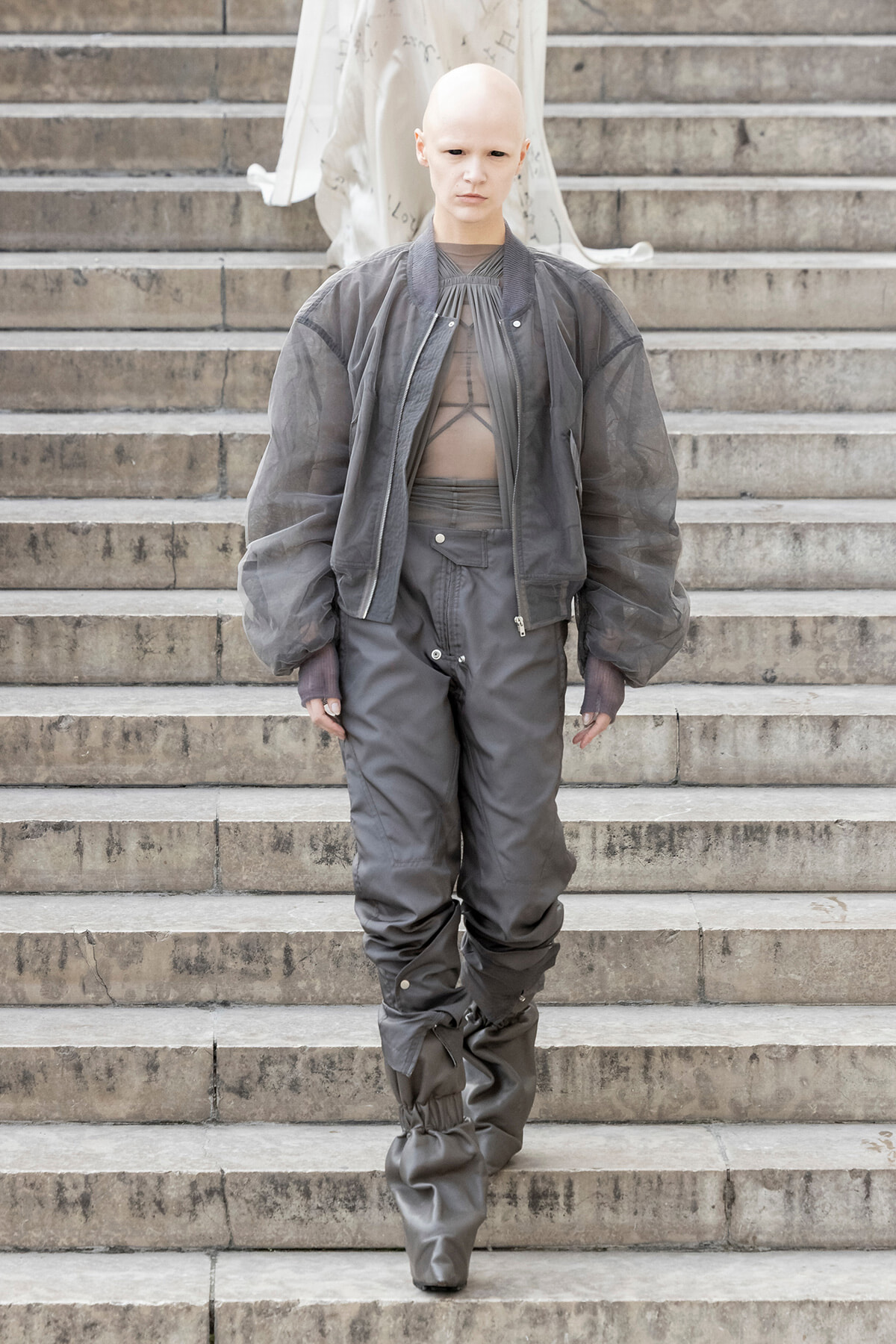 Bald model wearing a gray oversized bomber jacket and baggy gray pants walking down concrete stairs outside.