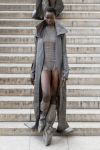 Model in a long gray trench and matching bodysuit descends stone steps, wearing knee-high sculptural boots.