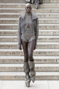 Model walking down concrete stairs in a monochrome gray athleisure look: oversized jacket, cropped top, ruched shorts, and tall slouch boots.