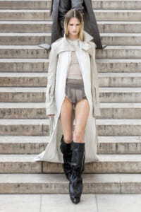 Model walking down stone stairs in a long light trench, sheer top and gray shorts, tall black boots, neutral-toned ensemble.