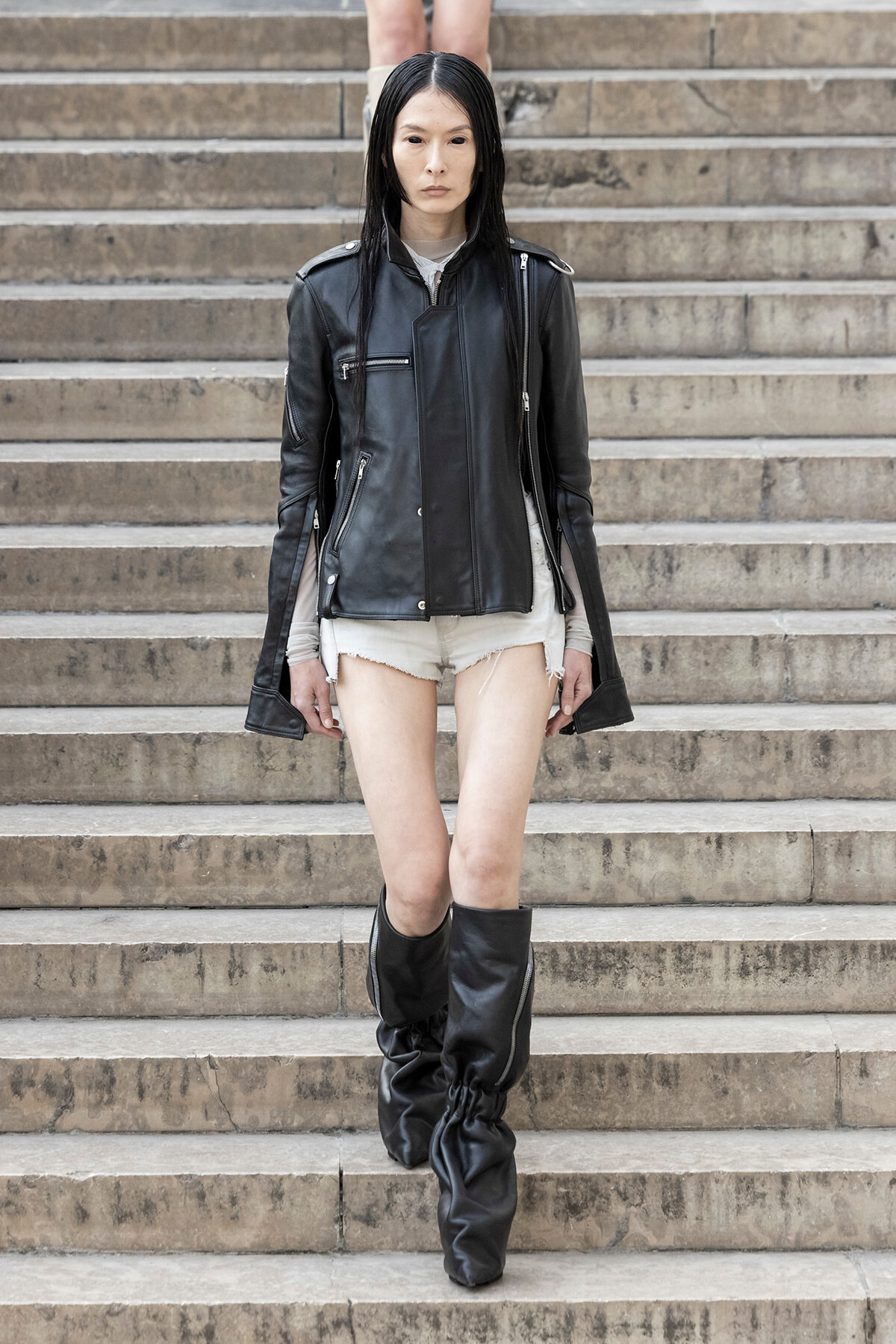 Woman wearing a black leather jacket, white top, and frayed beige shorts walking down stone stairs in tall black boots.