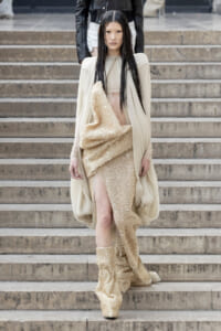 Model wearing a cream cape over a gold sequin dress with a high slit, walking down stone steps.