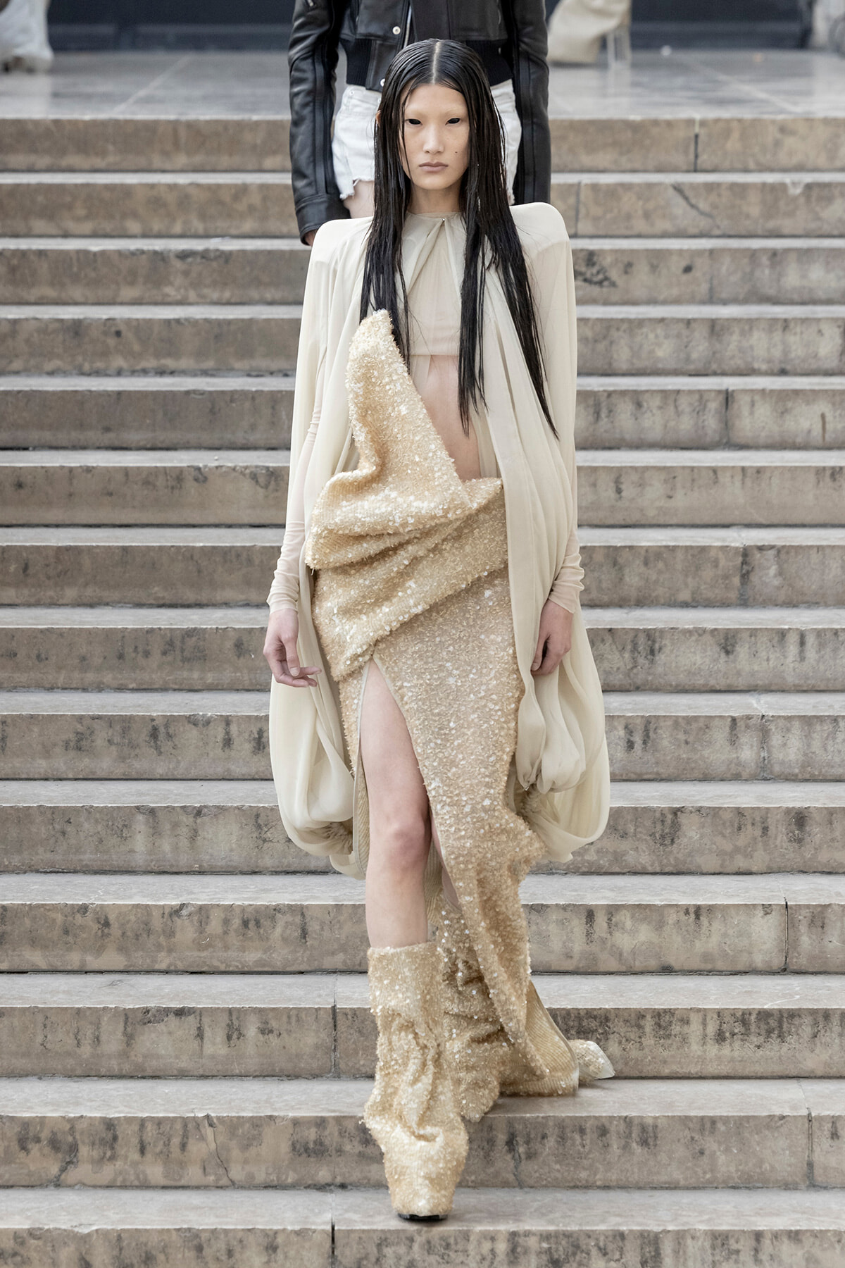 Model wearing a cream cape over a gold sequin dress with a high slit, walking down stone steps.