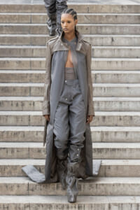Female model descending stone steps in a taupe trench coat over a gray jumpsuit with gathered, slouchy boots; serious expression.