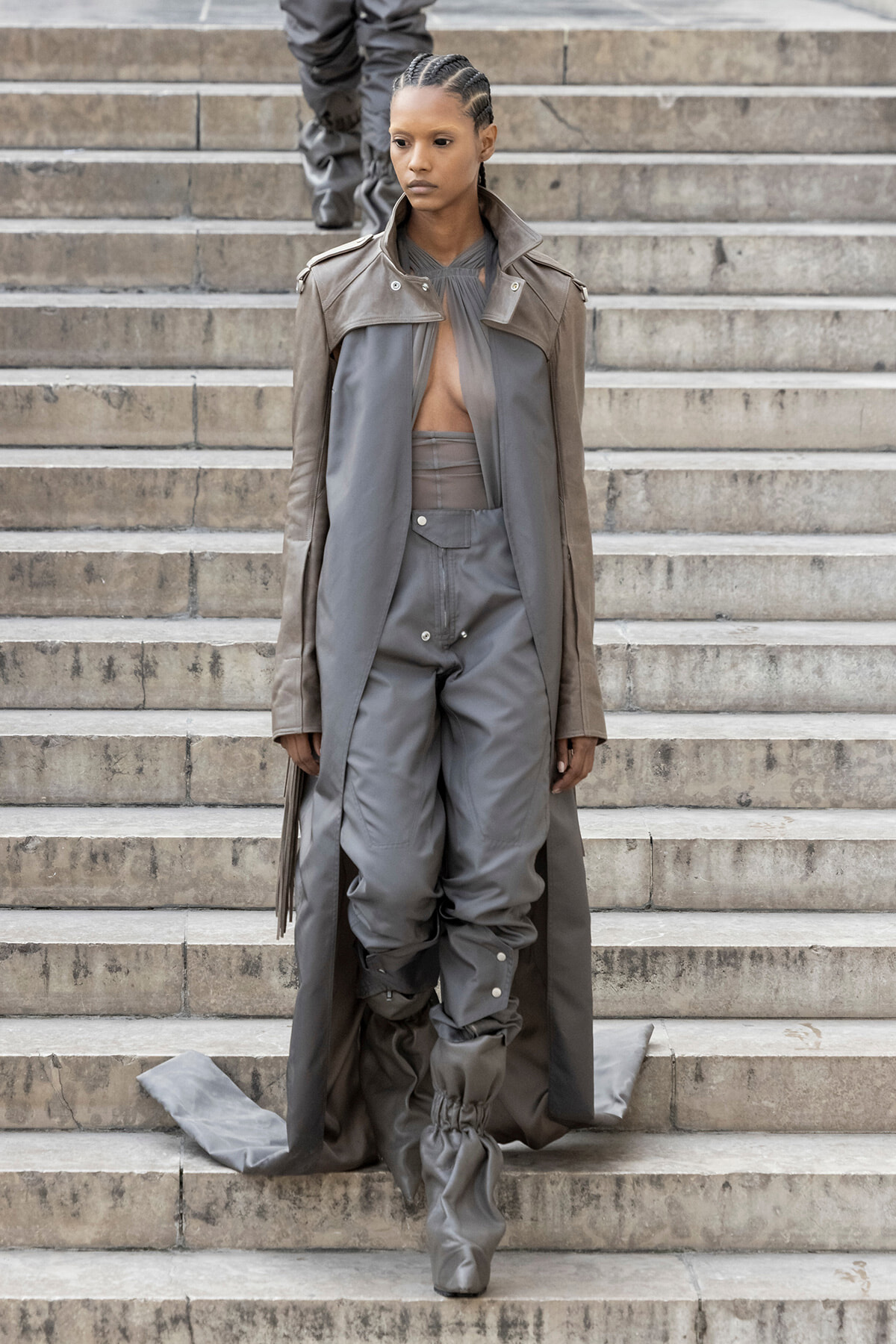 Female model descending stone steps in a taupe trench coat over a gray jumpsuit with gathered, slouchy boots; serious expression.