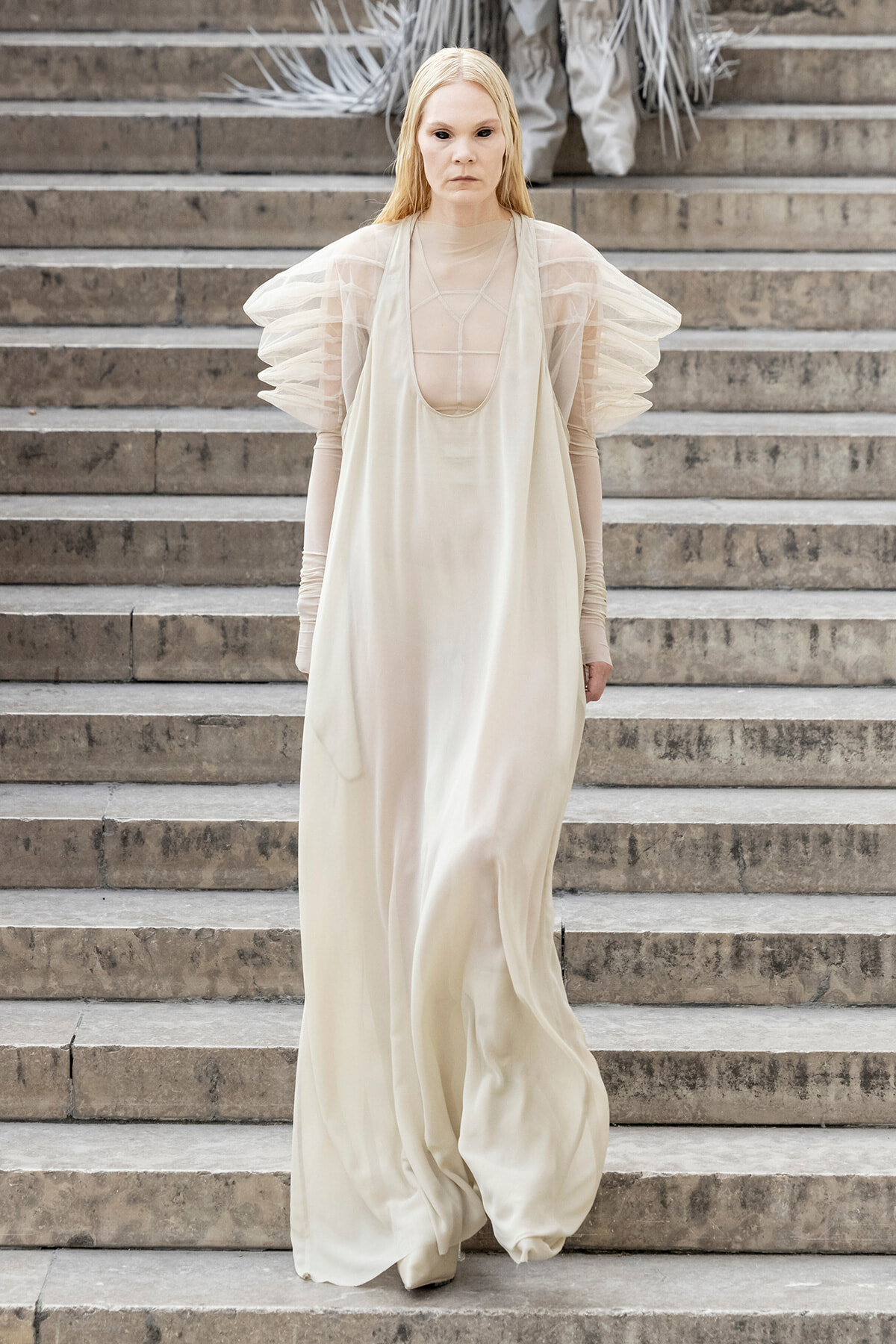 Model in an ivory, flowing gown with sheer puff sleeves walking down stone steps on a runway