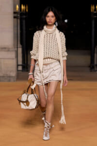 Model walks the runway in a beige crochet sweater, cream scarf, and matching shorts, carrying a tan-cream handbag with braided tassels.