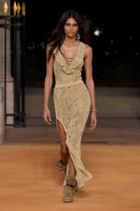 Tall model walking a beige lace maxi dress on a runway at night, with ruffled neckline and slit at the leg edge.
