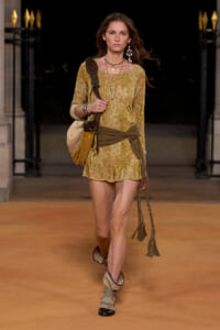 Model walking a runway in a mustard knit mini dress with a wrapped belt, tan fringe bag, and layered jewelry.