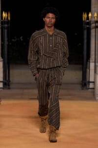 Male model walking a runway in a brown striped matching shirt and pants with tan suede boots.2) A male fashion model on a catwalk wearing a brown vertical-striped set and boots, hands in pockets.3) Runway model in brown striped loungewear-style shirt and wide-leg pants, tan suede boots.4) Model wearing brown pinstriped matching top and trousers with mid-calf suede boots at a fashion show.