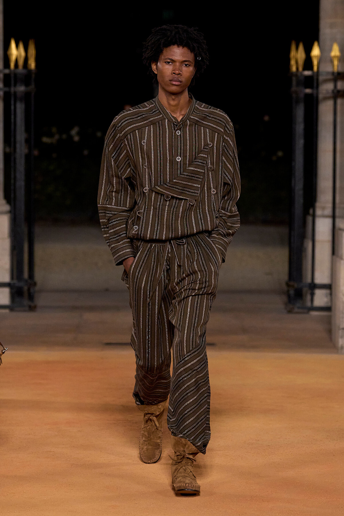 Male model walking a runway in a brown striped matching shirt and pants with tan suede boots.2) A male fashion model on a catwalk wearing a brown vertical-striped set and boots, hands in pockets.3) Runway model in brown striped loungewear-style shirt and wide-leg pants, tan suede boots.4) Model wearing brown pinstriped matching top and trousers with mid-calf suede boots at a fashion show.