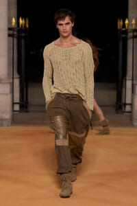 Male model walking runway in a beige knit sweater and brown patchwork pants with tan boots, posing mid-stride at dusk-lit venue.