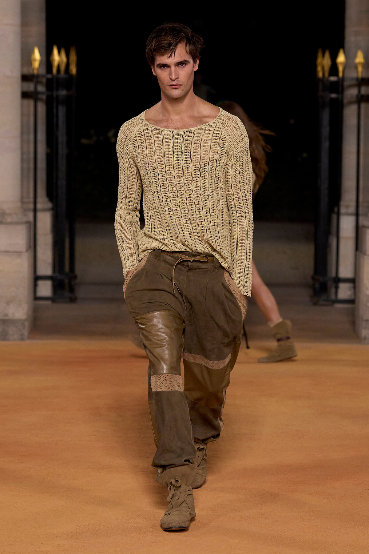 Male model walking runway in a beige knit sweater and brown patchwork pants with tan boots, posing mid-stride at dusk-lit venue.