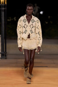 Model walking runway in a cream lace two-piece set with scalloped edges and geometric cutouts, beige suede boots.