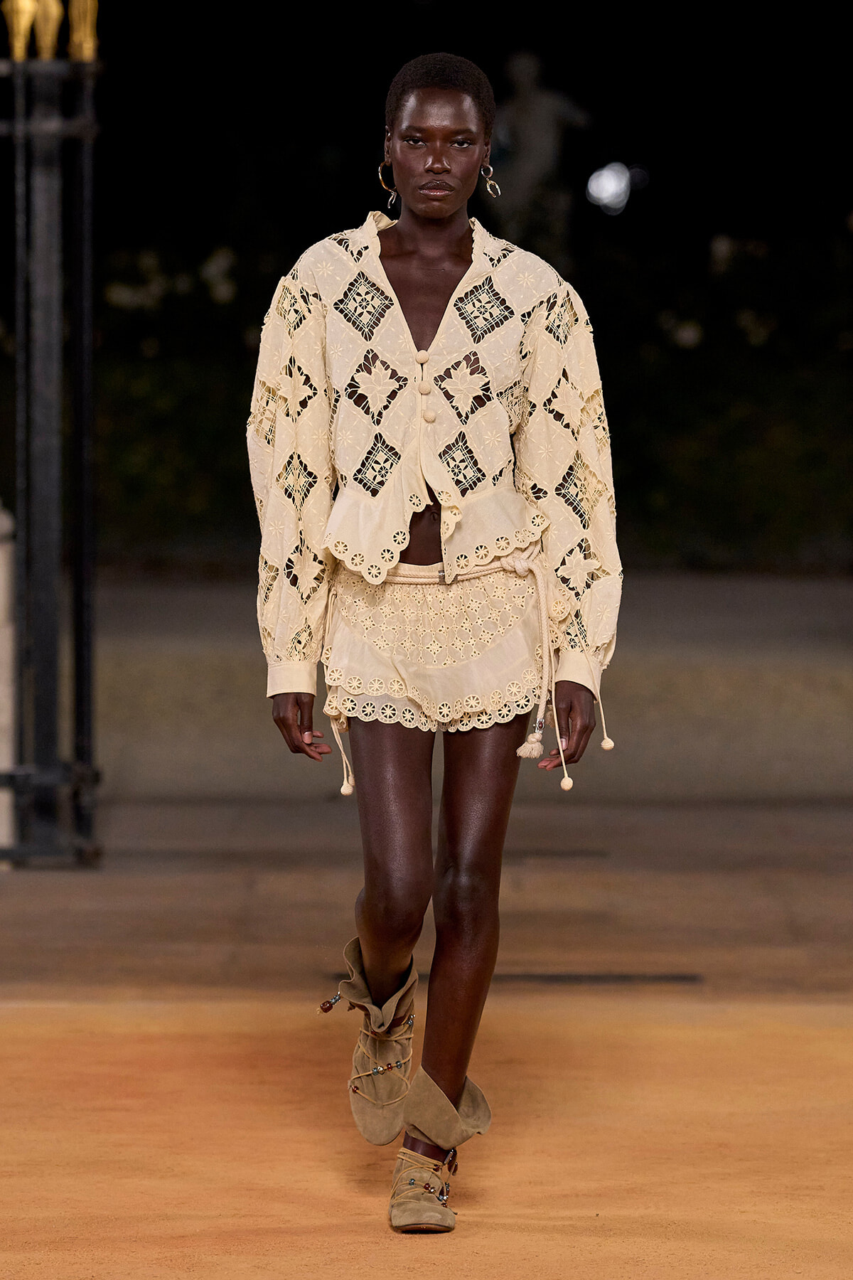 Model walking runway in a cream lace two-piece set with scalloped edges and geometric cutouts, beige suede boots.
