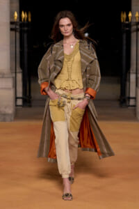 Runway model in earthy, layered look: striped coat, mustard lace-up top, and beige patchwork pants with orange lining.