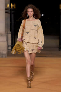 Model walking a runway in a loose beige knit sweater and matching shorts, carrying a tan shoulder bag with fringed boots.