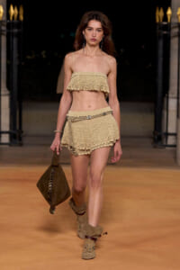 Model walking a runway in a beige crocheted two-piece outfit with a matching belt bag and tan boots, carrying a brown suede tote.