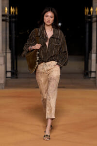 Model walks a runway wearing a dark striped oversized shirt tucked into light-brown tie-dye pants, with a brown shoulder bag and flat sandals.