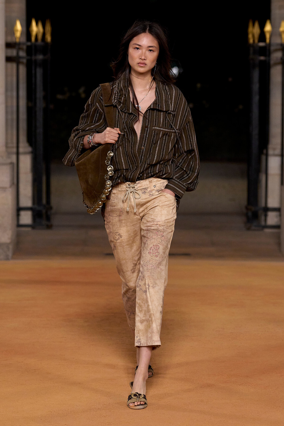 Model walks a runway wearing a dark striped oversized shirt tucked into light-brown tie-dye pants, with a brown shoulder bag and flat sandals.