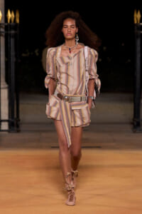 Model walking a runway at night in a pastel striped matching top and shorts with a wide belt and layered jewelry.