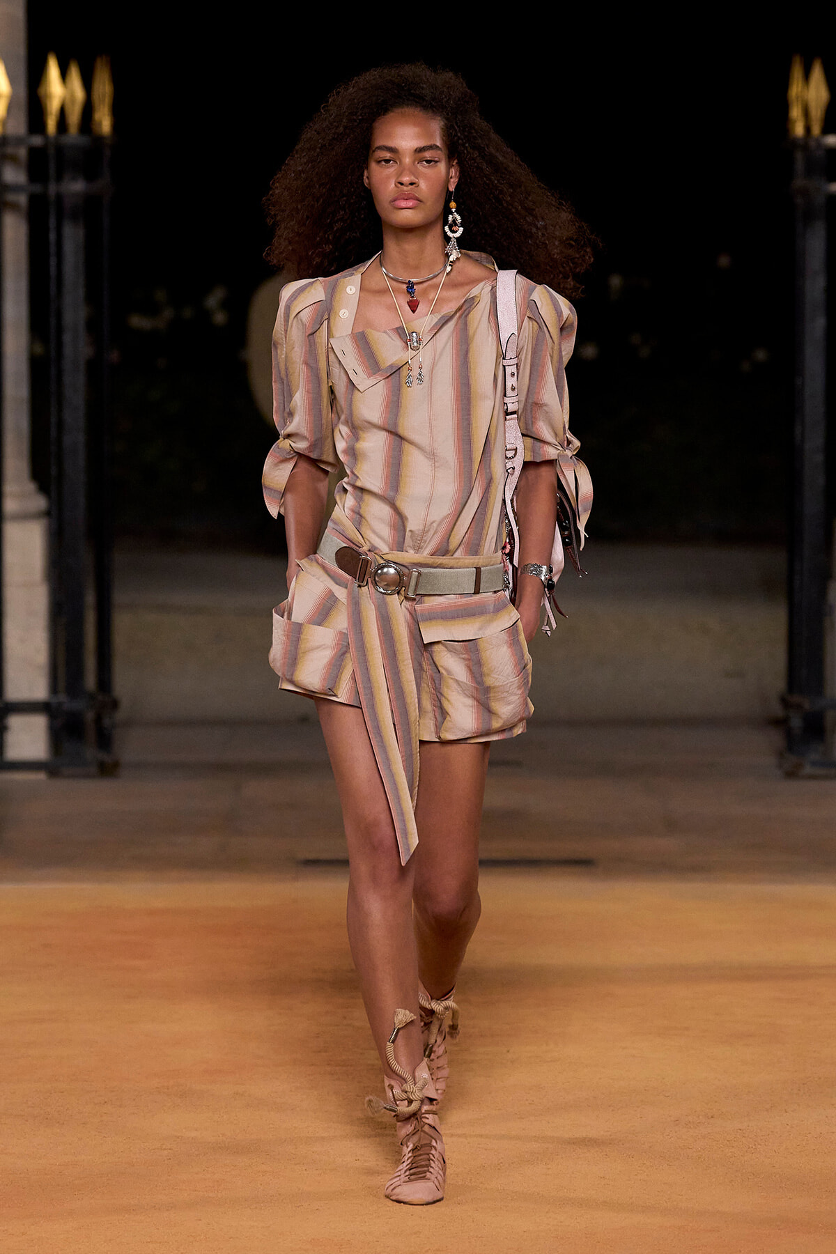 Model walking a runway at night in a pastel striped matching top and shorts with a wide belt and layered jewelry.